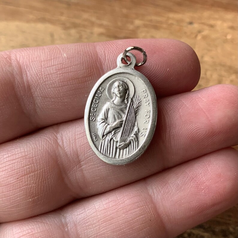 K&ouml;nnte beinhalten: Silberner ovaler Anh&auml;nger mit einer Darstellung der Heiligen C&auml;cilia, die eine Harfe und eine Palmenzweig h&auml;lt. Der Anh&auml;nger tr&auml;gt die Aufschrift "St. Cecilia" und "Pray for Us".