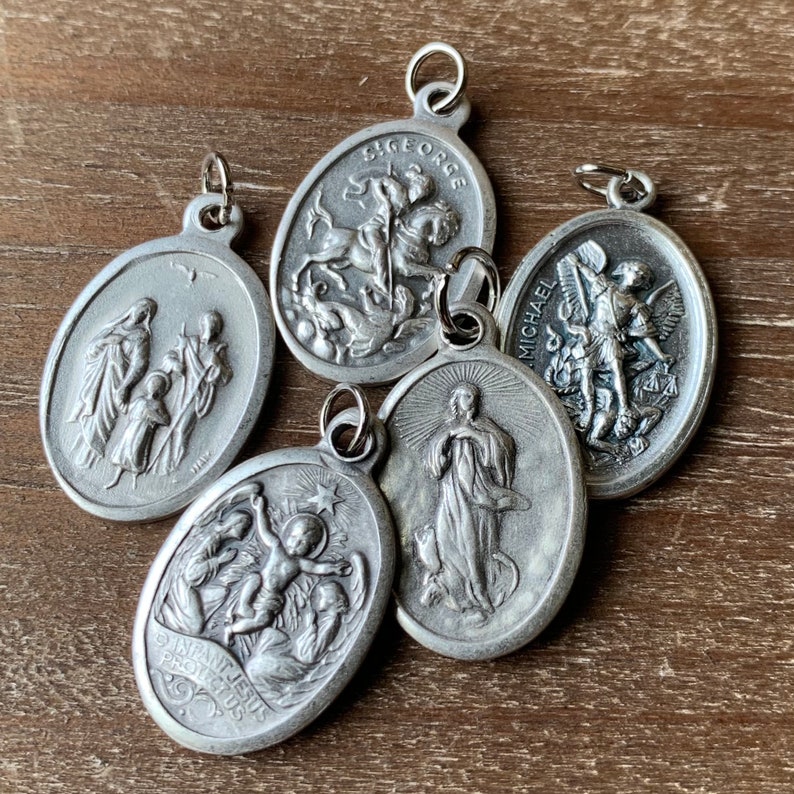 K&ouml;nnte beinhalten: F&uuml;nf silberne ovale religi&ouml;se Medaillons mit verschiedenen Bildern und Texten. Die Medaillons zeigen Bilder des Heiligen Georg, des Heiligen Michael, der Heiligen Familie, der Jungfrau Maria und des Jesuskindes. Der Text auf den Medaillons lautet "Heiliger Georg", "Heiliger Michael", "Heilige Familie", "Jesuskind" und "Sch&uuml;tze uns".
