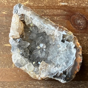 May include: A geode cut in half, revealing a sparkling interior of quartz crystals. The outer shell displays bands of gray, white, and brown. The geode sits on a wooden surface, showcasing its natural beauty.