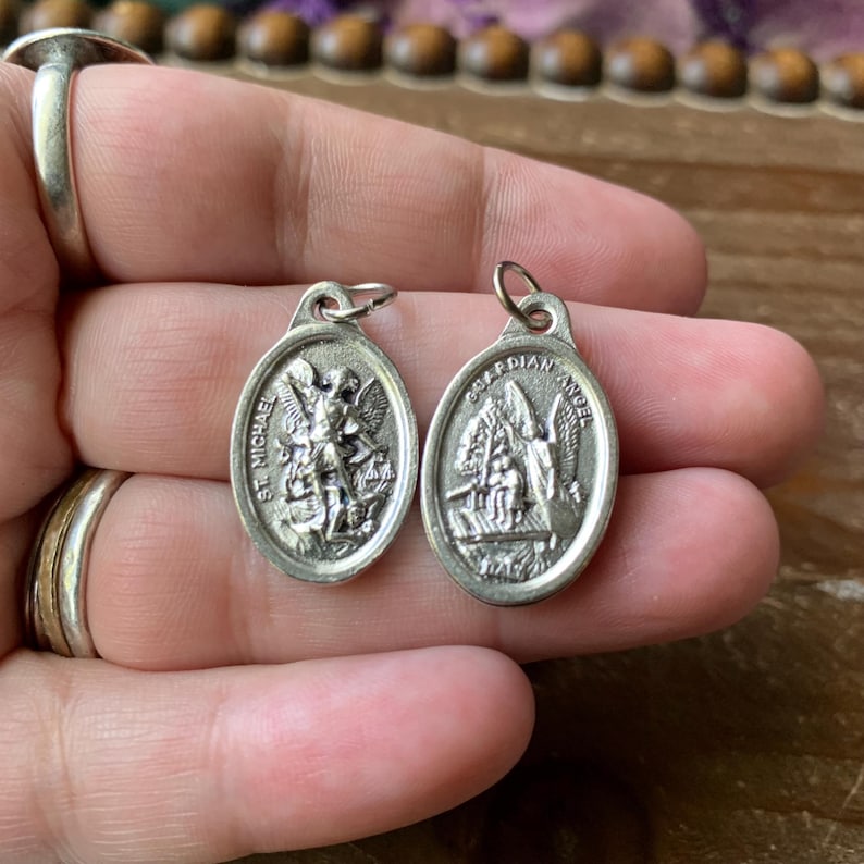 K&ouml;nnte beinhalten: Zwei silberne ovale Anh&auml;nger mit religi&ouml;sen Motiven. Ein Anh&auml;nger zeigt den Erzengel Michael mit dem Text "St. Michael" und der andere Anh&auml;nger zeigt einen Schutzengel mit dem Text "Guardian Angel".