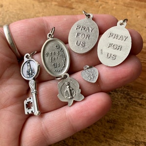 以下が含まれることがあります： 鍵、ハート、そして「Pray for Us」と刻まれた4つの楕円形のチャームを含む、6つの銀色の宗教的なチャームのセットです。