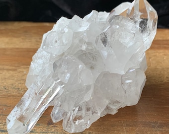 May include: A cluster of clear quartz crystals, showcasing a variety of pointed formations. The crystals are transparent, reflecting light and revealing internal structures. The quartz sits on a wooden surface, with a dark background.