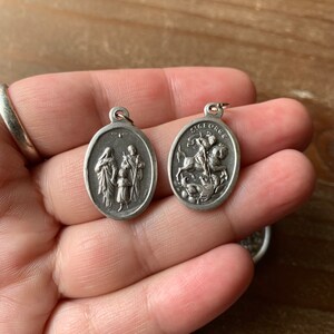 K&ouml;nnte beinhalten: Zwei silberne ovale Anh&auml;nger mit religi&ouml;sen Motiven. Ein Anh&auml;nger zeigt eine Darstellung des Heiligen Georg, der einen Drachen t&ouml;tet, mit der Inschrift "S. GEORGE". Der andere Anh&auml;nger zeigt eine dreik&ouml;pfige Familie, wahrscheinlich die Heilige Familie.