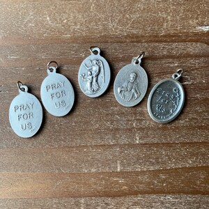 K&ouml;nnte beinhalten: F&uuml;nf silberne ovale Anh&auml;nger mit religi&ouml;sen Motiven. Jeder Anh&auml;nger tr&auml;gt den Text "Pray for Us" und das Wort "Italy".