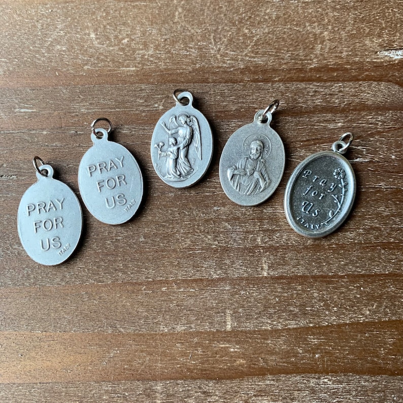 K&ouml;nnte beinhalten: F&uuml;nf silberne ovale Anh&auml;nger mit religi&ouml;sen Motiven. Jeder Anh&auml;nger tr&auml;gt den Text "Pray for Us" und das Wort "Italy".