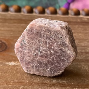 May include: A rough, pink ruby crystal with a hexagonal shape. The crystal has a natural, unpolished finish and is a beautiful example of this gemstone.
