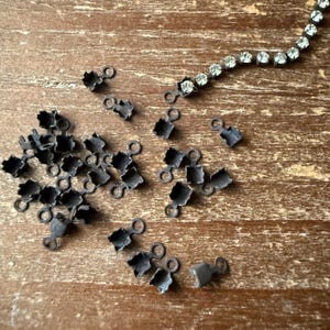 May include: A collection of small, dark metal jewelry findings and a rhinestone chain on a wooden surface. The findings have a square shape with a loop and are scattered across the wood. The rhinestone chain is silver and curved.