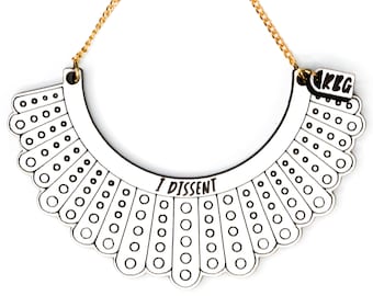 May include: A white wooden necklace with a gold chain, featuring a black outline of a judge's collar and the text "I Dissent". The necklace has a small tag with the initials "RBG" in black.