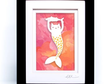 May include: A framed artwork featuring a white cat-mermaid hybrid holding a fish above its head. The creature has a yellow polka-dot tail and is set against a vibrant pink and orange watercolor background. The piece is enclosed in a black frame.