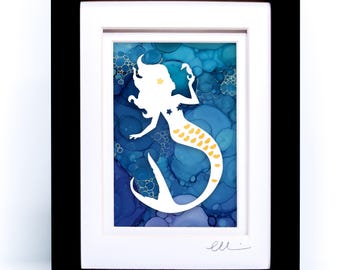 May include: A framed artwork featuring a white silhouette of a mermaid against a blue alcohol ink background. The mermaid has gold accents on her tail and a small gold star in her hair. The piece is enclosed in a black frame with a white mat.