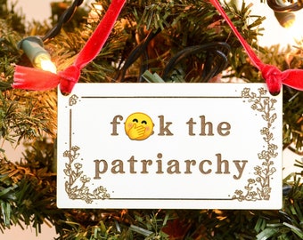 May include: A wooden Christmas ornament with a red ribbon and the text "Fok the patriarchy" with a yellow emoji face with a hand over its mouth.