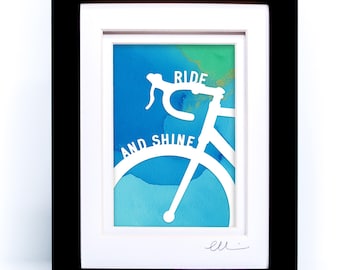 May include: A framed art print featuring a white silhouette of bicycle handlebars and a partial frame against a blue and green watercolor background. The text RIDE AND SHINE is printed in white across the bicycle frame. The frame is black.