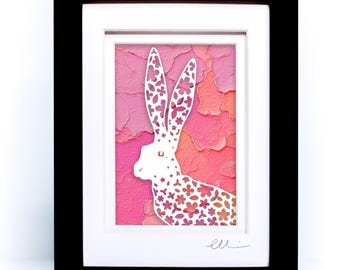 May include: A framed artwork featuring a white paper-cut silhouette of a hare with intricate floral cutouts. The hare is set against a textured background of layered pink, coral, and orange paper. The piece is enclosed in a black frame with a white mat.