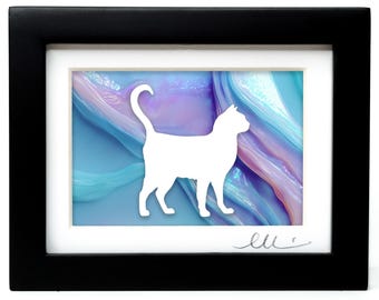 May include: A framed artwork featuring a solid white silhouette of a standing cat centered against a background of swirling blue, purple, and pink colors. The piece is enclosed in a rectangular black frame with a white mat and a signature in the corner.