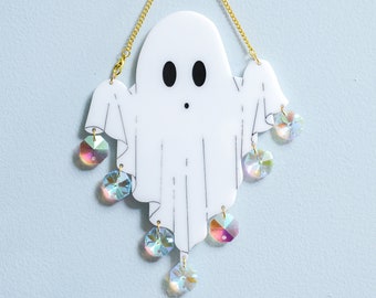May include: A white ghost-shaped ornament with a gold chain hanging from the top. The ghost has black eyes and a small mouth. The ornament is decorated with iridescent crystals hanging from the bottom.