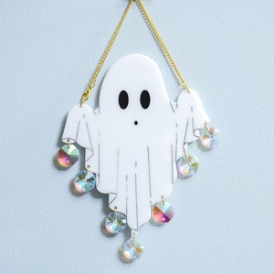 May include: A white ghost-shaped ornament with a gold chain hanging from the top. The ghost has black eyes and a small mouth. The ornament is decorated with iridescent crystals hanging from the bottom.