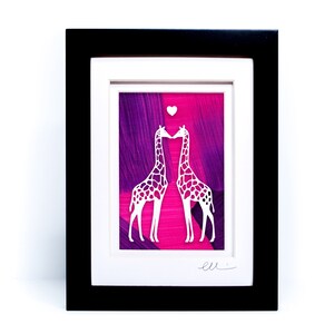 May include: A framed paper cut art piece featuring two white giraffes kissing with a small heart above them. The background is a pink and purple abstract painting.