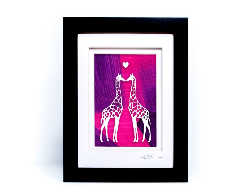 May include: A framed paper cut art piece featuring two white giraffes kissing with a small heart above them. The background is a pink and purple abstract painting.