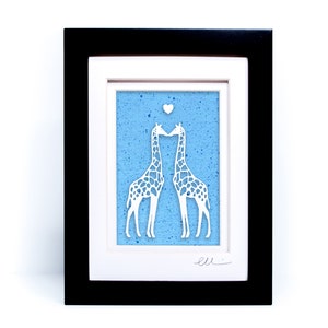 May include: A framed paper cut-out art piece depicting two white giraffes kissing on a blue background with white speckles. A small white heart is above the giraffes. The piece is signed "ell."
