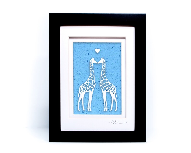 May include: A framed paper cut-out art piece depicting two white giraffes kissing on a blue background with white speckles. A small white heart is above the giraffes. The piece is signed "ell."