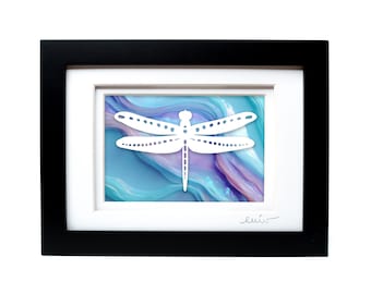 May include: A framed artwork featuring a white paper-cut dragonfly silhouette centered over a background of swirling blue, purple, and teal colors. The piece is enclosed in a black rectangular frame with a white mat and a signature in the corner.
