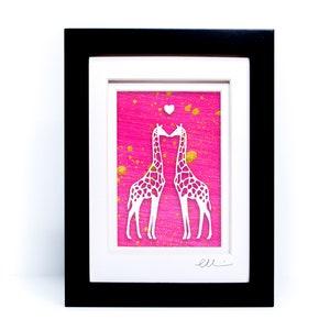May include: A framed paper cut-out art piece featuring two white giraffes kissing on a pink background with gold splatter paint. The giraffes are facing each other and their necks are intertwined. A small white heart is above their heads. The artwork is signed "ell"