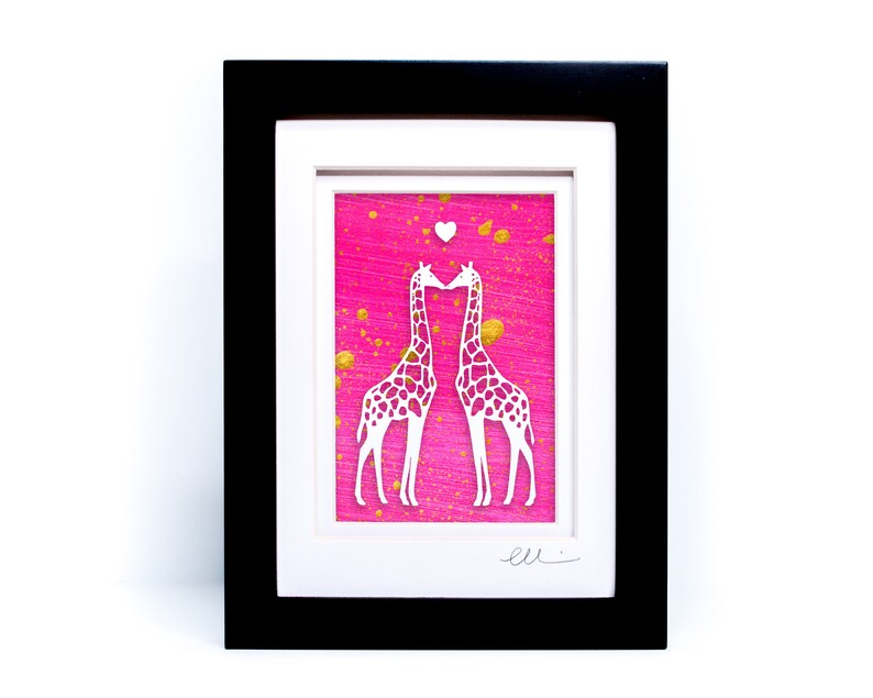 May include: A framed paper cut-out art piece featuring two white giraffes kissing on a pink background with gold splatter paint. The giraffes are facing each other and their necks are intertwined. A small white heart is above their heads. The artwork is signed "ell"
