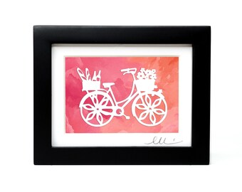 May include: A framed artwork featuring a white paper-cut silhouette of a bicycle with a front basket of flowers and a rear basket containing baguettes and a bottle, set against a vibrant pink and orange watercolor background with a signature.