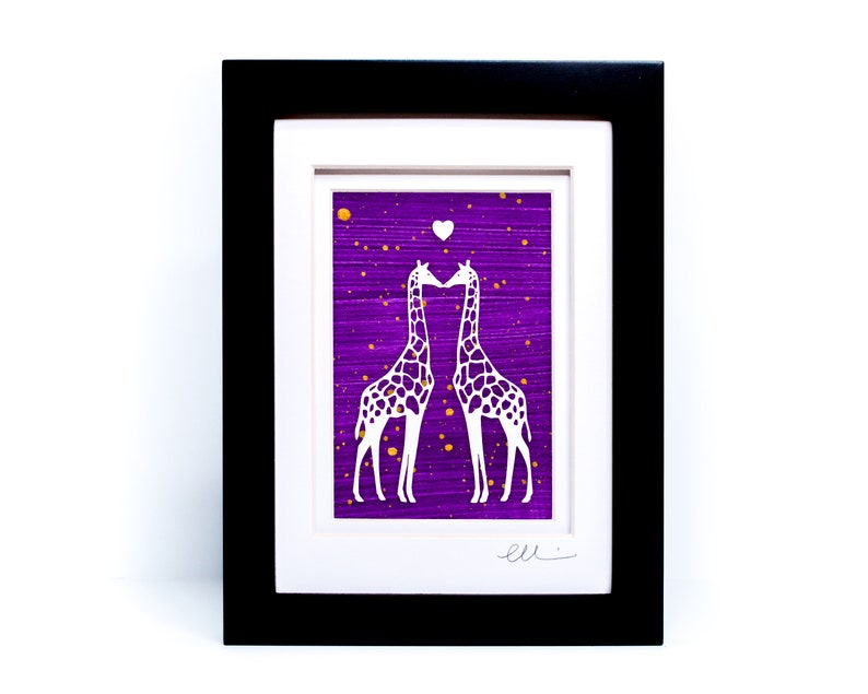May include: Framed paper cut art featuring two white giraffes kissing on a purple background with gold glitter. The giraffes are silhouetted against the purple background and have a small white heart above them. The artwork is signed "ell"