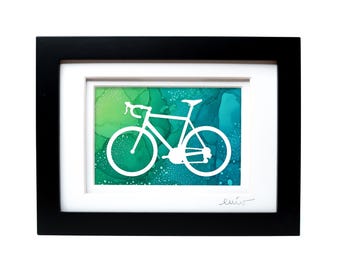 May include: A framed artwork featuring a white silhouette of a road bicycle centered against a vibrant, abstract background of blended green and teal ink washes. The piece is enclosed in a simple black frame with a white mat and a signature at the bottom.
