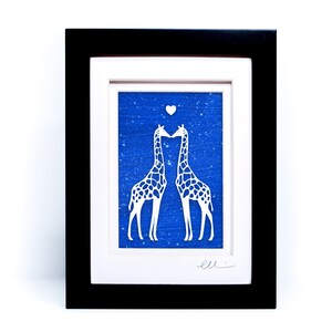 May include: A framed paper cut art piece depicting two giraffes kissing with a heart above them. The giraffes are white against a blue background with white speckles.