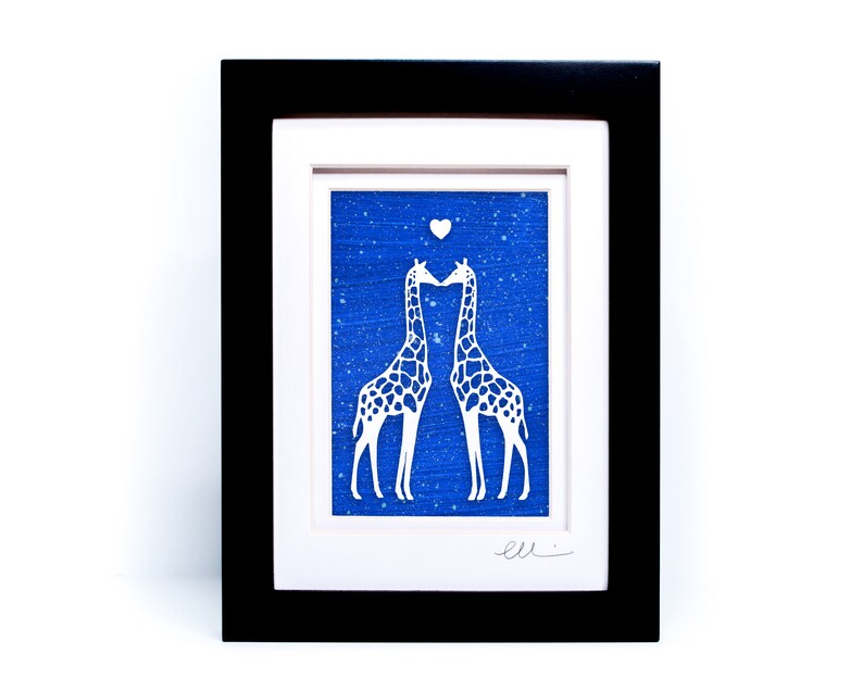 May include: A framed paper cut art piece depicting two giraffes kissing with a heart above them. The giraffes are white against a blue background with white speckles.
