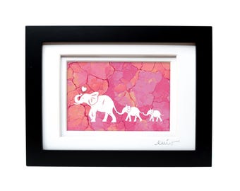 May include: A framed artwork featuring a white silhouette of an adult elephant and two smaller elephants walking in a line against a textured pink and orange background. The adult elephant holds a small white heart above its trunk. The frame is black.