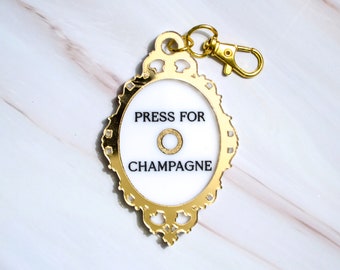 May include: Gold-colored keychain with a white circle in the center that says "Press for Champagne".