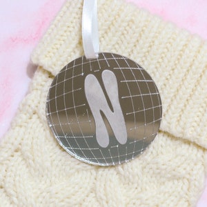 May include: A silver ornament with a grid pattern and the letter N in a stylized font. The ornament is hanging from a white ribbon on a cream knitted fabric.