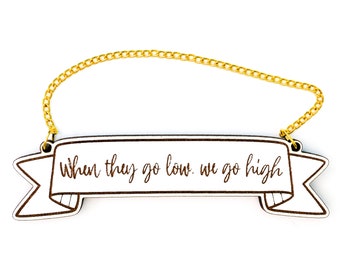 May include: Wooden banner with a gold chain hanging from the top. The banner has the text "When they go low, we go high" written in brown cursive.