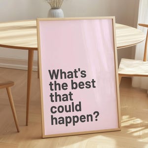 Puede incluir: Un póster enmarcado con un fondo rosa claro y el texto "What's the best that could happen?" en negro. El póster está apoyado contra una pared en una habitación con muebles de madera y un jarrón con flores secas.