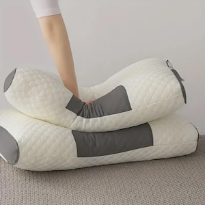 以下が含まれることがあります： テクスチャ加工された表面とグレーのアクセントが施された2つの白い枕。1つの枕は湾曲しており、もう1つは長方形です。枕は首をサポートするように設計されており、柔らかい素材で作られています。