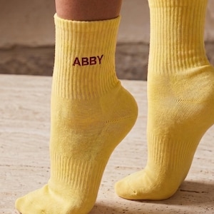 Op de afbeelding: Een paar felgele kuit-sokken met een geribbelde textuur. De naam ABBY is geborduurd in donkerrood garen aan de buitenkant van de linkersok. De sokken hebben een effen kleurontwerp met een eenvoudige gebreide constructie.