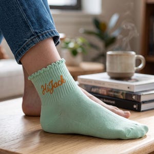 May include: A light mint green ankle sock with a ruffled top edge and the word "HiJack" embroidered in orange cursive script on the side. The sock features a ribbed cuff design and is made of a smooth, solid-colored fabric material.