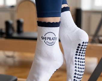 Może przedstawiać: Białe skarpetki z ciemnoniebieskim, falbankowym ściągaczem i okrągłym logo z napisem SHJ PILATES STUDIO. Podeszwy posiadają wzór małych, ciemnoniebieskich silikonowych wypustek w kształcie serc, zapewniających przyczepność podczas ćwiczeń.