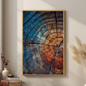 May include: A framed wall art print featuring a close-up of a tree stump. The artwork showcases concentric rings in shades of blue, orange, and brown, with a gold-colored frame. The piece is displayed in a room with neutral tones and natural light.