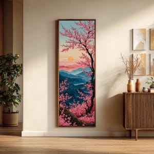 May include: A framed vertical artwork showing a Japanese landscape. A cherry blossom tree with pink flowers is in the foreground. The background features blue mountains, a setting sun, and a light blue sky. The artwork is displayed on a beige wall.