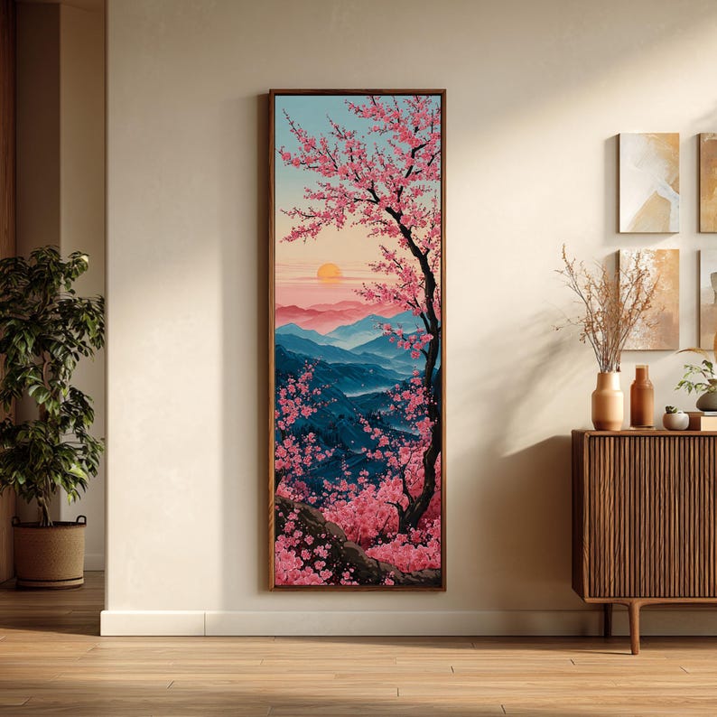 May include: A framed vertical artwork showing a Japanese landscape. A cherry blossom tree with pink flowers is in the foreground. The background features blue mountains, a setting sun, and a light blue sky. The artwork is displayed on a beige wall.