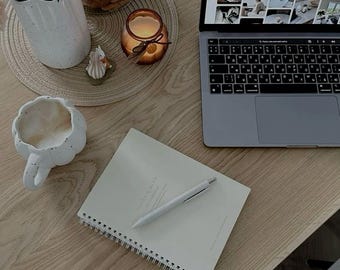 May include: A light wood table with a laptop, notebook, pen, flowers, and a candle. The laptop displays a website. A white mug and a small decorative item are also on the table. The notebook has the words "Flora & Mind" printed on it.