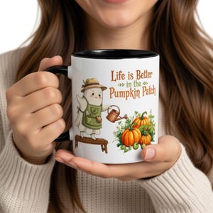 May include: A white ceramic mug with a black handle and rim, featuring a cartoon ghost wearing a straw hat and green apron, standing on a wooden bench while watering orange pumpkins. The text reads, Life is Better in the Pumpkin Patch.