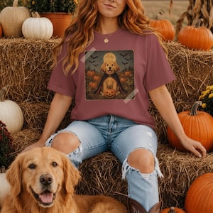 Puede incluir: Una camiseta color rosa polvoriento con un estampado gráfico de un golden retriever con capa de vampiro, sentado en un campo de calabazas bajo una luna llena con murciélagos. El diseño incluye una pequeña calabaza a los pies del perro.