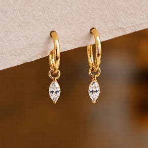 May include: A pair of gold hoop earrings featuring a dangling marquise-cut clear gemstone charm. The earrings are constructed with a polished gold-tone metal finish and a secure hoop closure, showcasing a simple and elegant design for everyday wear.
