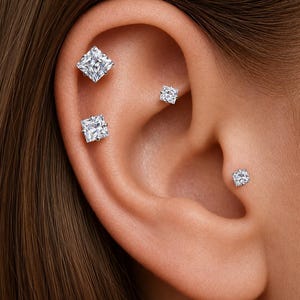 May include: Four silver-toned stud earrings featuring clear, square-cut faceted stones. The earrings are arranged in the ear, with two larger square studs in the upper cartilage, one smaller square stud in the conch, and one small square stud in the tragus.
