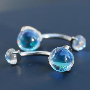 May include: Two pairs of silver earrings with blue and clear glass orb details. The earrings are presented on a dark blue surface, with light reflecting off the glass. The earrings have a modern, minimalist design.